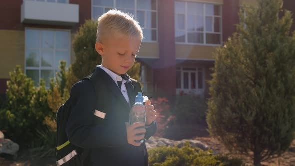 A Small Schoolboy with a Backpack Drinks Water From a Bottle in the School Yard