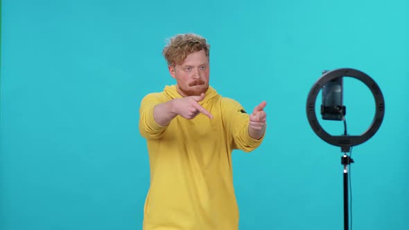 Young Man Influencer in Yellow Hoodie Dances on a Blue Light Background and Takes a Selfie Video on