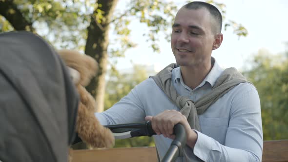 Close-up Portrait of Happy Young Father Playing Teddy Bear in Sunny Park with Infant in Baby