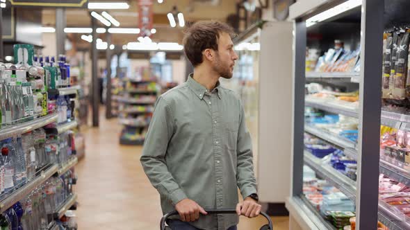 Happy Man in Shirt Walking with a Cart Through the Supermarket Choosing Groceries