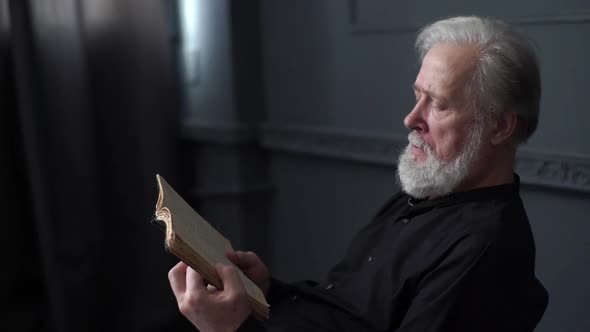 Tracking Shot of Thoughtful Grayhaired Senior Aged Man Reading Older Paper Book in Dark Library Room