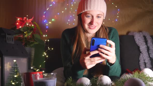 Young Happy Woman Sitting on the Sofa in Christmas Decorated Living Room Using Mobile Phone. 