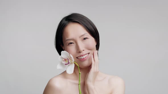 Middle Aged Asian Lady Holding Orchid Touching Face Gray Background