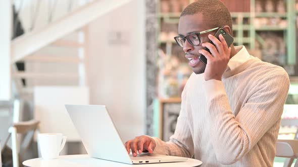 African Man with Laptop Talking on Smartphone in Cafe