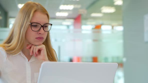 A Young Girl with Glasses in a Spacious Office Working on a Laptop.