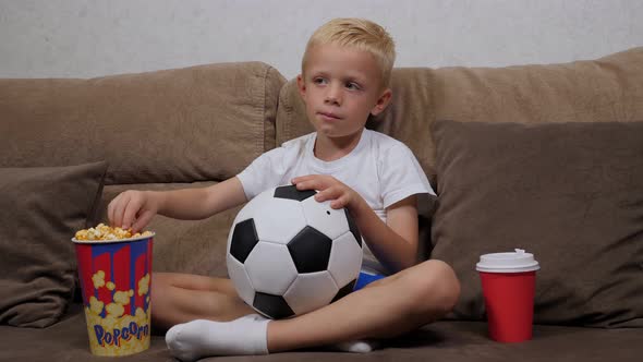 A Little Boy is Watching a Football Match on TV Sitting on Couch with Popcorn