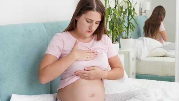 Young Pregnant Woman in Pajamas Sitting on Bed and Examining Her Breast for Cancer