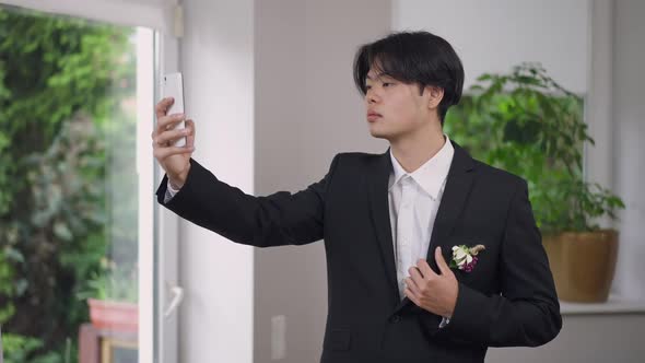 Joyful Young Asian Groom in Elegant Wedding Suit Taking Selfie on Smartphone Indoors