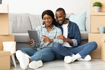  Among Cardboard Boxes After Moving To New Flat, Smiling African American Couple Using Tab Computer For Online Shopping Or Browsing Internet