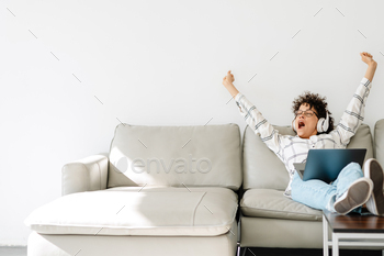 Young woman in headphones yawning and using laptop while sitting on couch / Photodune, Misc
