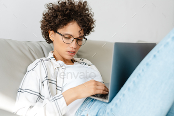 Young curly woman using laptop while sitting on couch at home / Photodune, Misc