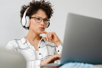 Young curly woman in headphones using laptop while sitting on couch / Photodune, Misc
