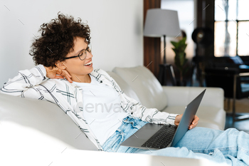 Young curly woman smiling and using laptop while sitting on couch / Photodune, Misc