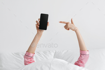 f young unrecognizable female under blanket holds smartphone, shows finger at blank screen of gadget on white wall background in bedroom, copy space