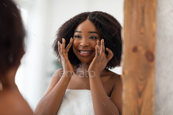 checking first wrinkles, admiring soft silky skin after face cream at home. African American woman looking at her reflection after skincare treatment