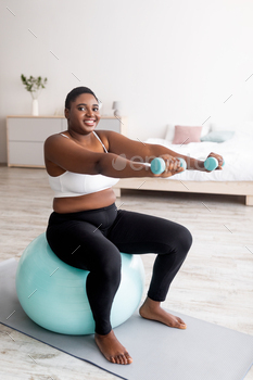 Cheerful plus size Afro woman exercising with dumbbells on fitness ball at home, full length / Photodune, Misc