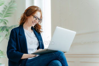 Professional female employer enjoys working process, sits crossed legs with laptop device