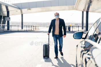 Business traveler pulls suitcase from modern airport terminal, walks to car, wears medical mask / Photodune, Misc