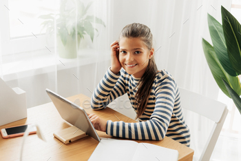 Smiling schoolgirl in a striped sweater sits at the table, does homework on the tablet / Photodune, Misc