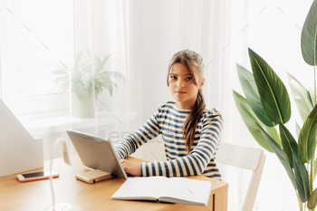 A serious schoolgirl in a striped sweater is sitting at a table, doing her homework on her tablet / Photodune, Misc