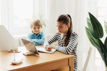 A schoolgirl, sitting at a table with her younger brother, performs an assignment on a tablet / Photodune, Misc