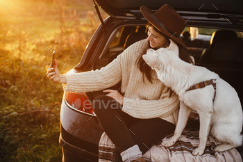 Autumn road trip with pet. Stylish hipster woman taking selfie photo with cute dog in car trunk / Photodune, Misc