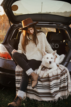 Stylish woman in hat and sweater sitting and hugging cute dog in car trunk in evening autumn field / Photodune, Misc
