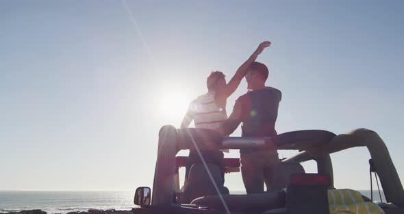 Happy caucasian gay male couple standing in car raising arms and embracing on sunny day at the beach