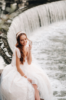 An elegant bride in a white dress, gloves and bare feet is sitting near a waterfall in the Park / Photodune, Misc