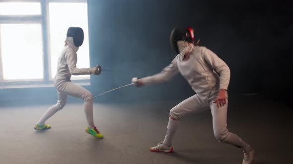 Two Young Women Having an Intense Training in a Fencing Duel in the Smoky Studio