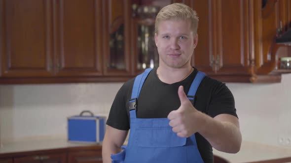 Portrait of Young Caucasian Handyman in Workrobe Standing at Kitchen and Showing Thumb Up. Handsome