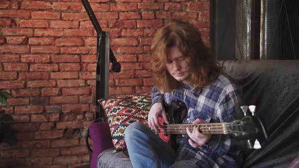 Portrait of Young Man Musician with Long Red Hair Playing Bass Guitar at Home Sitting on Sofa