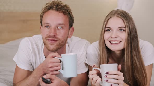 Smiling Young Couple Relaxing and Watching TV with Tea Cups at Home