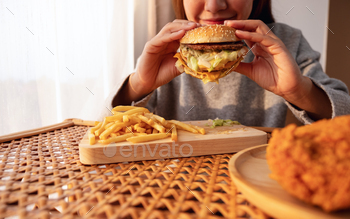 自宅のテーブルでハンバーガーとフライドチキンとフライドポテトを持って食べている女性 / Photodune, Misc