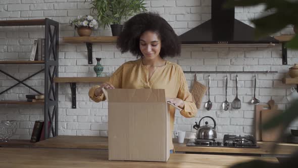 Happy Cheerful Frican American Girl Gets the Delivery Box. Happy Box Openning in a Kitchen