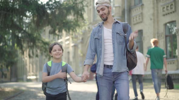 Portrait of Positive Caucasian Brothers Walking Along Schoolyard in Sunlight. Smiling Young Man and