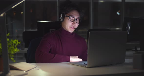 Asian Businessman in Headset Working with Computer Late at Night in Client Support Service