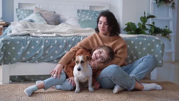 Young Mother and Daughter in Sweaters Sit on the Floor on a Rug with a Dog Jack Russell and