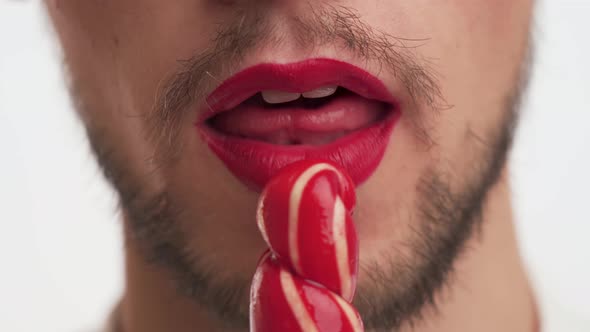 Pretty smiling man with makeup - red lipstick on lips open mouth wide, lick sweet sugar lollipop