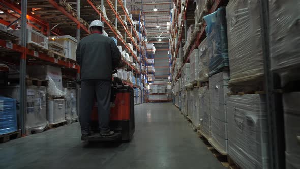 The Worker Rides on the Square Between the Shelves Filled with Boxes of Goods