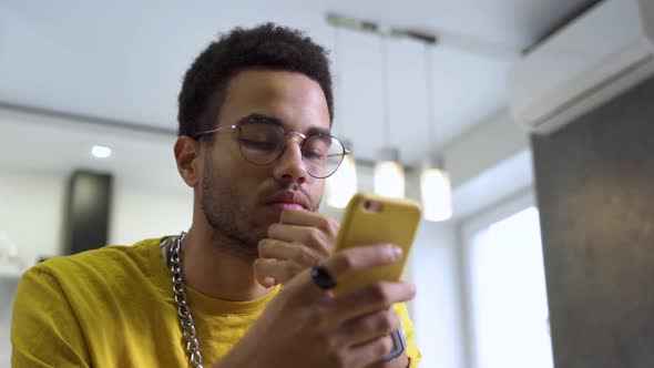 A Young Man Wearing Glasses Reads Messages on a Smartphone at Home