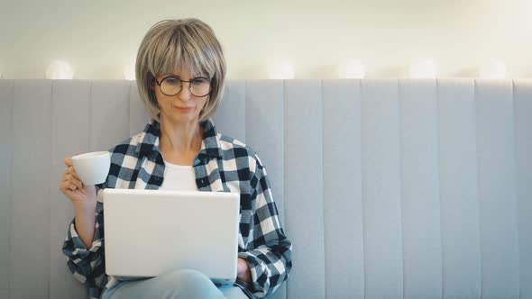 An Elderly Beautiful Woman Works with Interest at a Laptop and Drinks Coffee From a Cup Sitting at