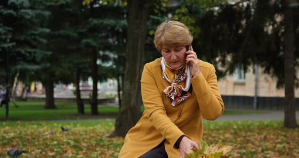 Senior Lady in a Yellow Jacket and a Handkerchief Around His Neck Sitting on a Park Bench Talking on