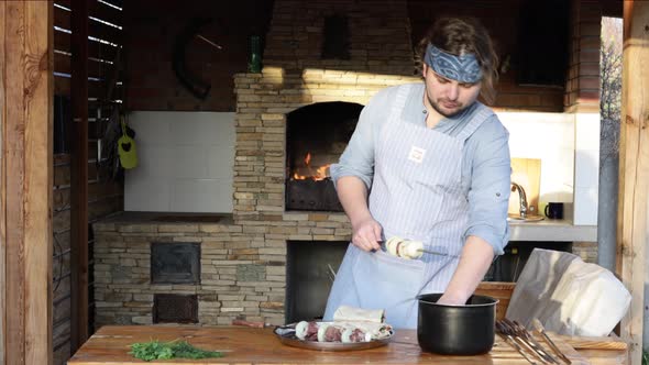 A Man in an Apron Strings Pieces of Meat on a Skewer and Makes a Kebab From It Near the Fireplace