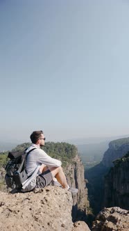 Traveler Guy with Glasses Sits on a Cliff and Looks at a Picturesque View