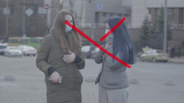 Two Young Caucasian Women in Protective Face Masks Talking Outdoors As Prohibition Cross Appearing