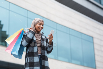 ping in city outdoor. Happy cute millennial islamic black female in hijab with multicolored purchases bags typing on phone near shop window of mall