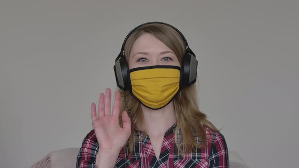 Portrait of Joyful Young Woman in Yellow Face Mask and Headphones Greeting Friends or Colleagues in