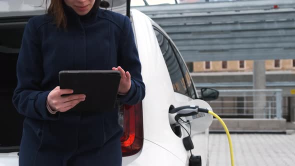 Business Woman Standing Near Charging Electric Car or EV Car and Using Tablet in the Street