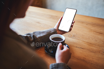 a woman holding black mobile phone with blank screen while drinking coffee on wooden table / Photodune, Misc